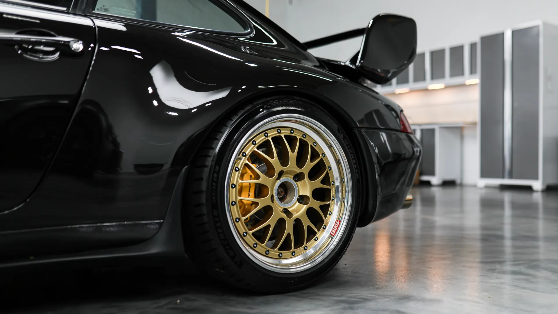 Close-up of a black sports car's rear with gold aftermarket wheels in a clean garage.
