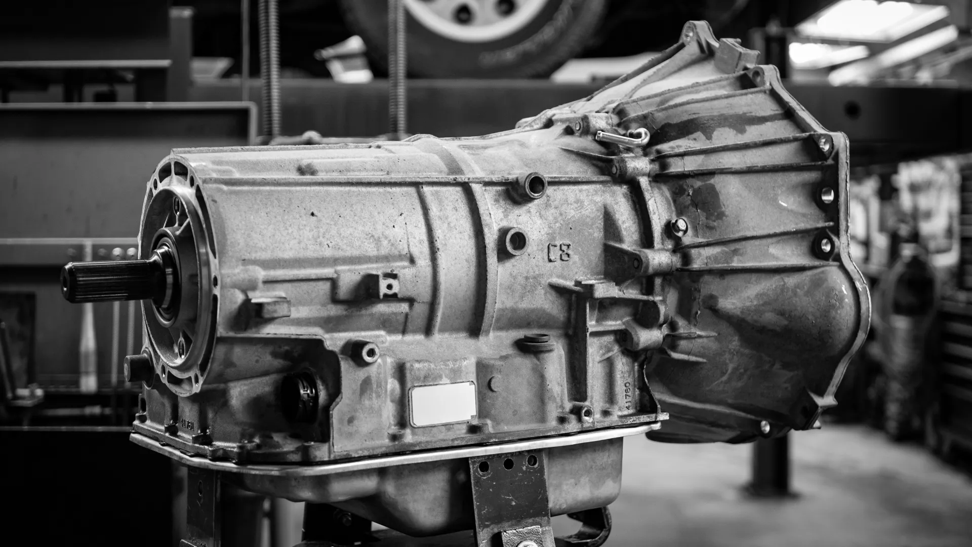 Black and white image of an automotive transmission on a stand in a workshop with a tire and tools in the background