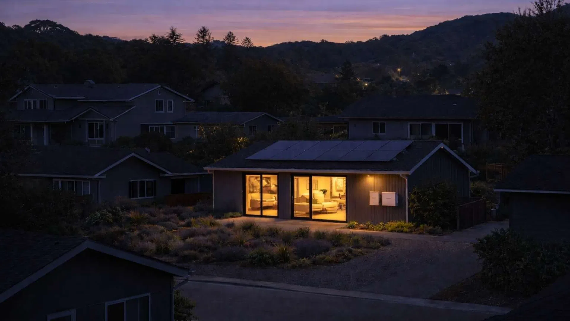 Nighttime view of a modern house with warm interior lighting and solar panels on the roof in a quiet neighborhood.