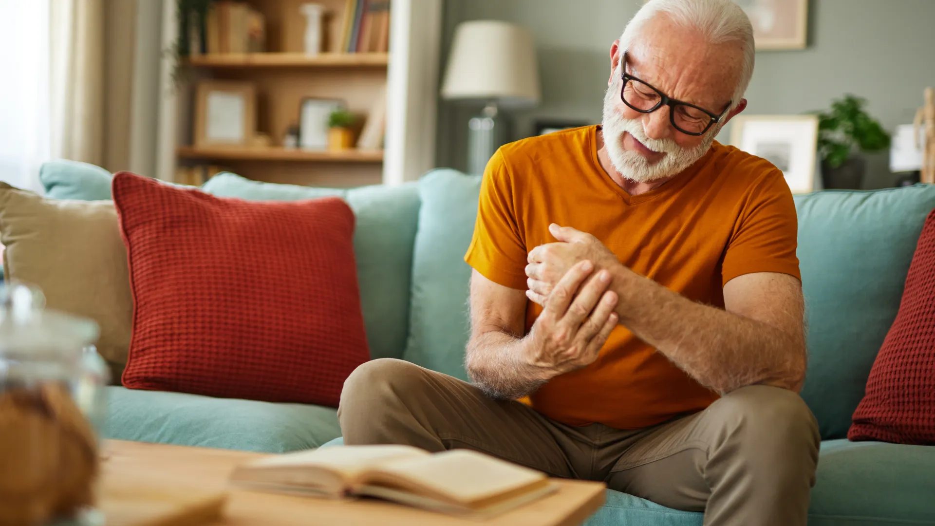 Elderly man with glasses sitting on couch, holding wrist in pain, showing discomfort indoors.