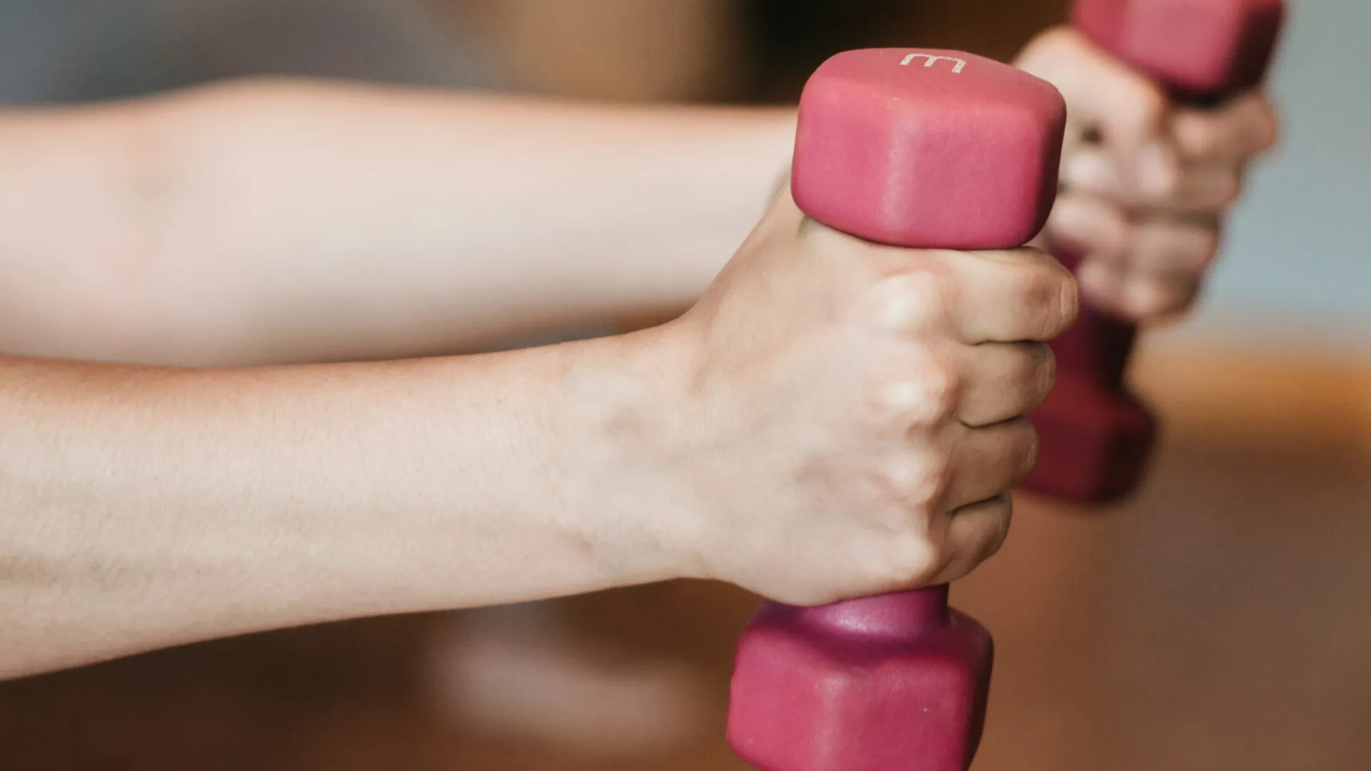 Close-up of hands holding pink dumbbells during a workout with blurred background person exercising indoors