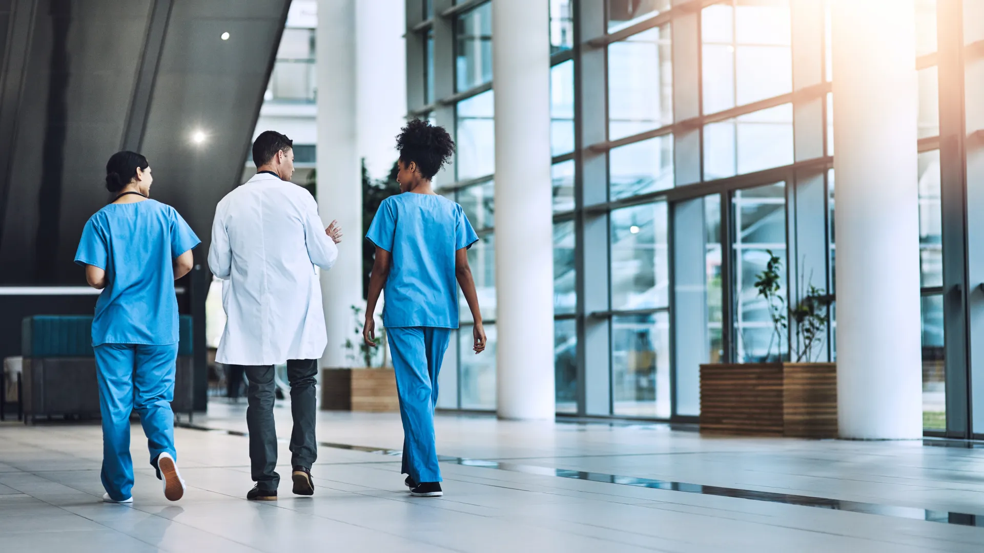 Three medical professionals walking and talking in a modern hospital corridor with glass walls.