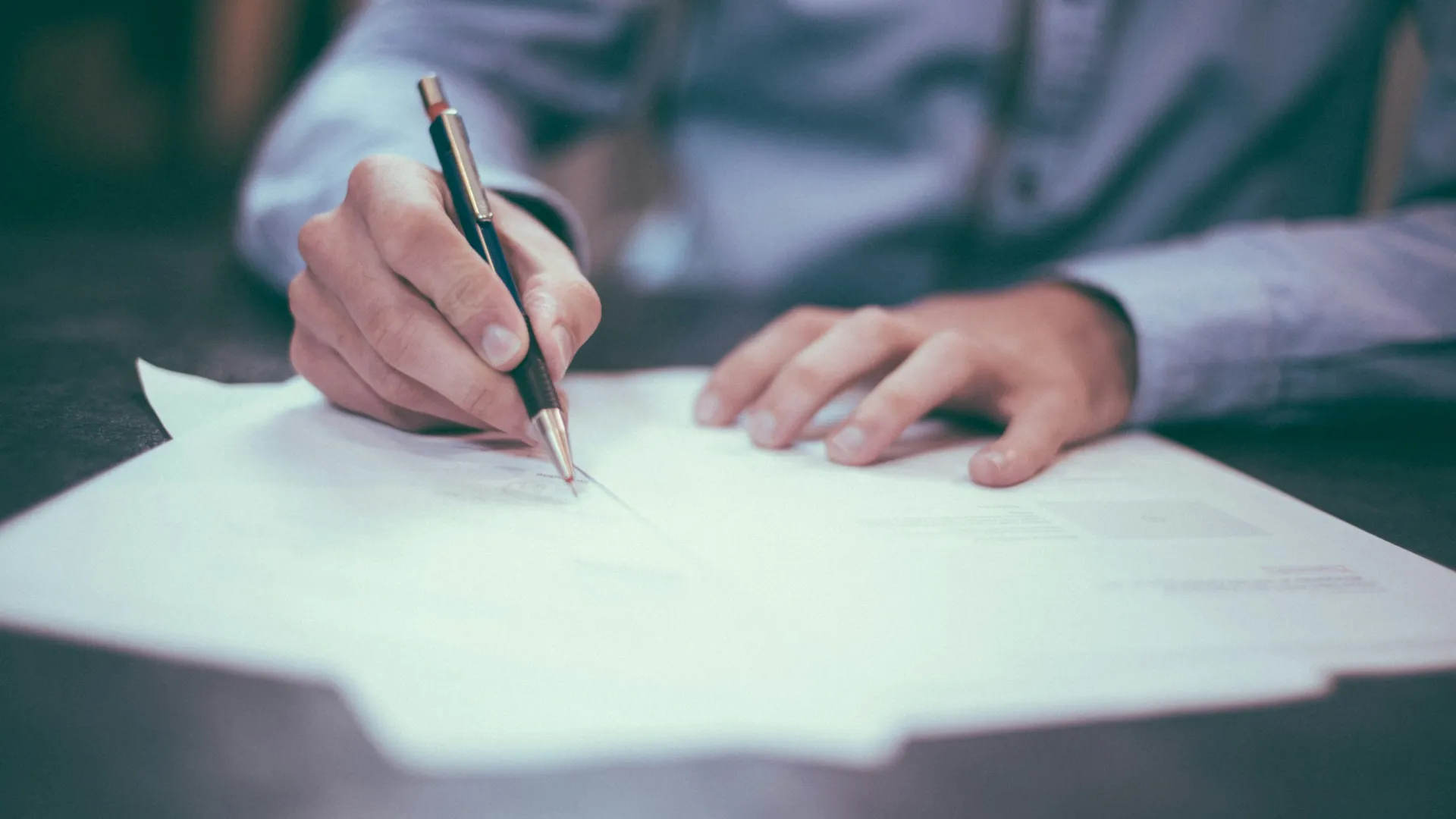 Person writing on a document with a pen on a desk, wearing a light blue long-sleeve shirt.