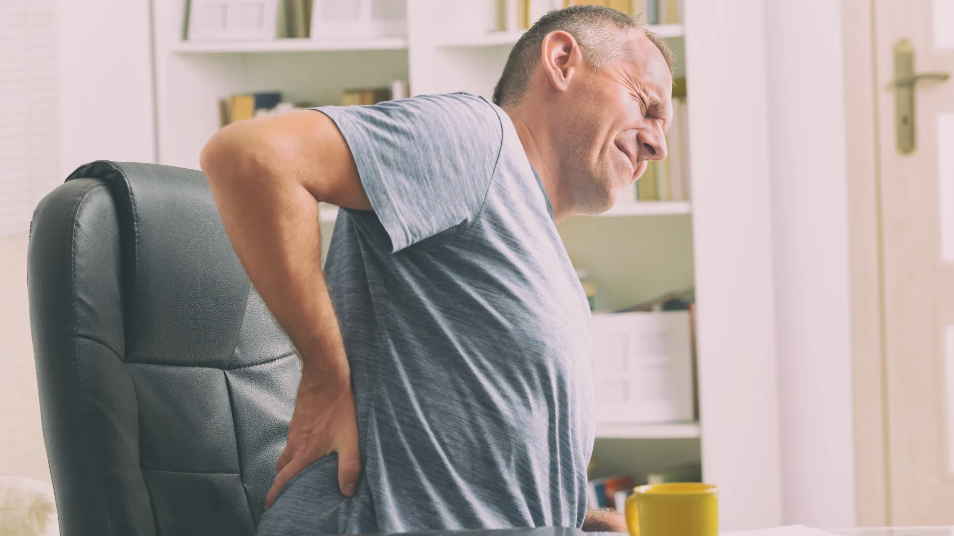 Man sitting in office chair holding lower back in pain, indicating discomfort or backache during work.