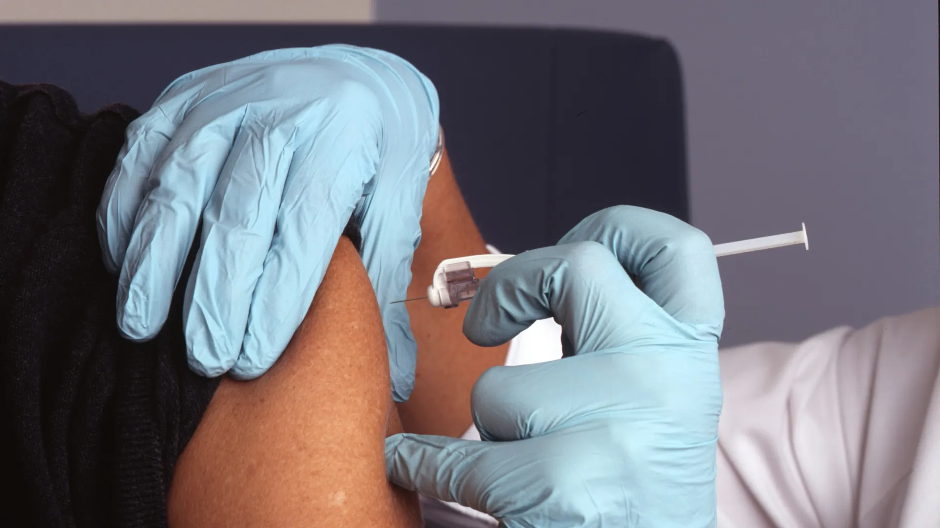 Healthcare worker wearing blue gloves administering a vaccine injection into a patient's upper arm.