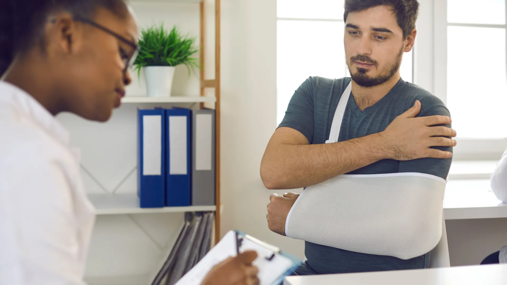 Man with arm in a sling discussing injury with female doctor taking notes in a medical office.