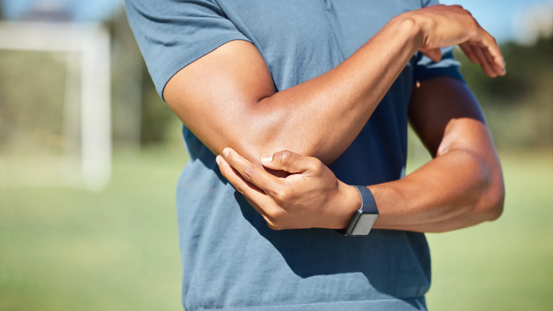 Man in blue shirt holding his elbow outdoors in a park or sports field during daytime