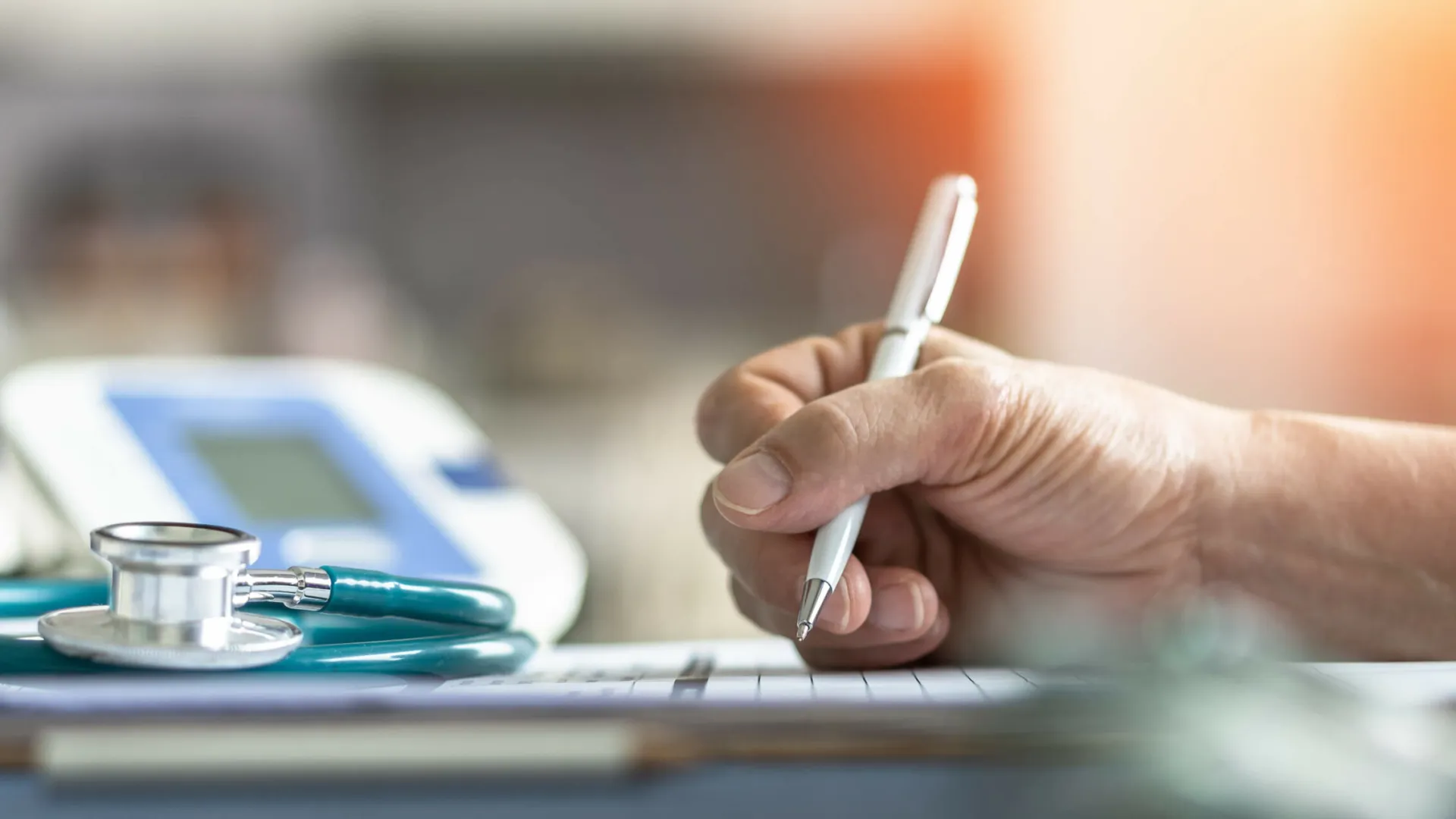Doctor writing notes with stethoscope and blood pressure monitor on a desk in bright room