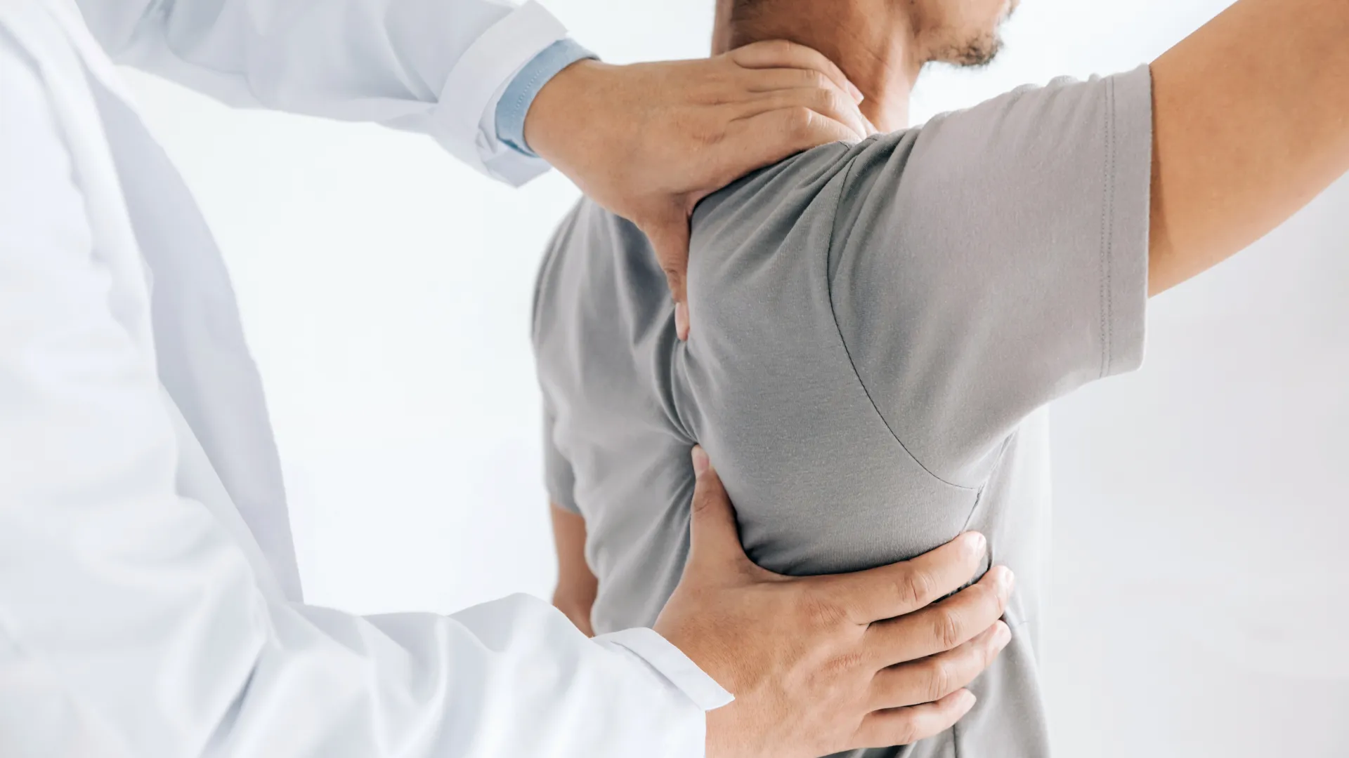 Doctor examining male patient's back and shoulder for pain or mobility issues in a clinical setting.