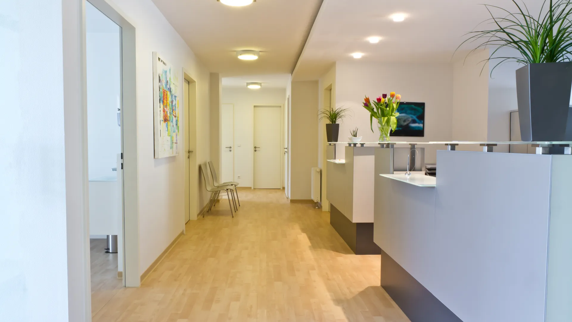 Modern medical office hallway with light wood flooring, white walls, a reception desk, and flower decorations.