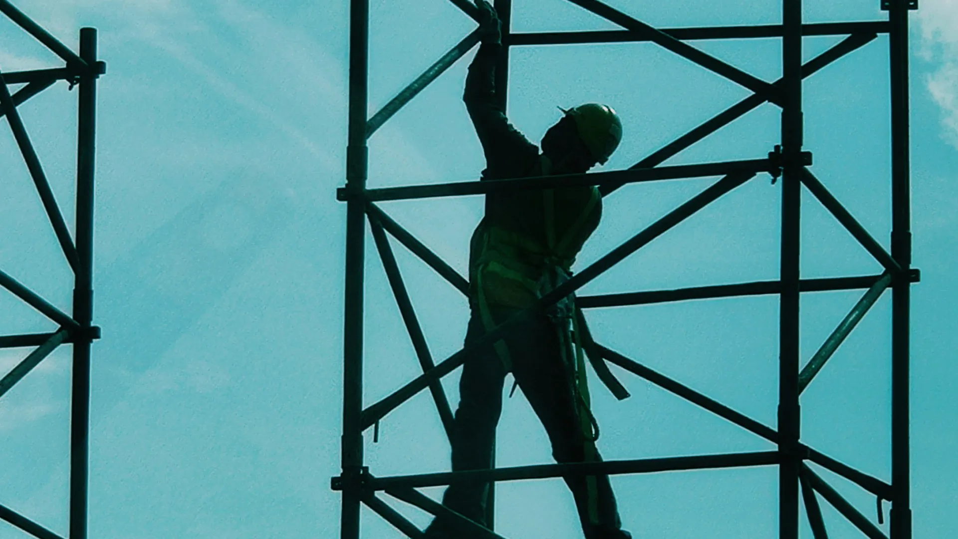 Silhouetted construction worker wearing helmet on scaffolding against a bright blue sky with clouds.