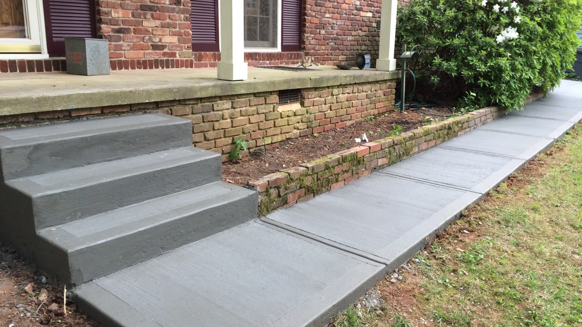 Freshly poured gray concrete stairs and walkway alongside a brick house with a garden bed and green shrub.