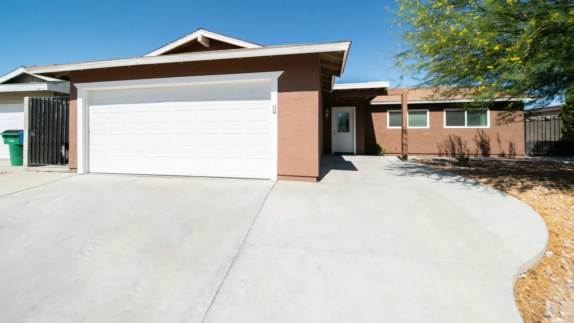 Single-story brown house with white garage door, concrete driveway, and sparse desert landscaping under clear blue sky.