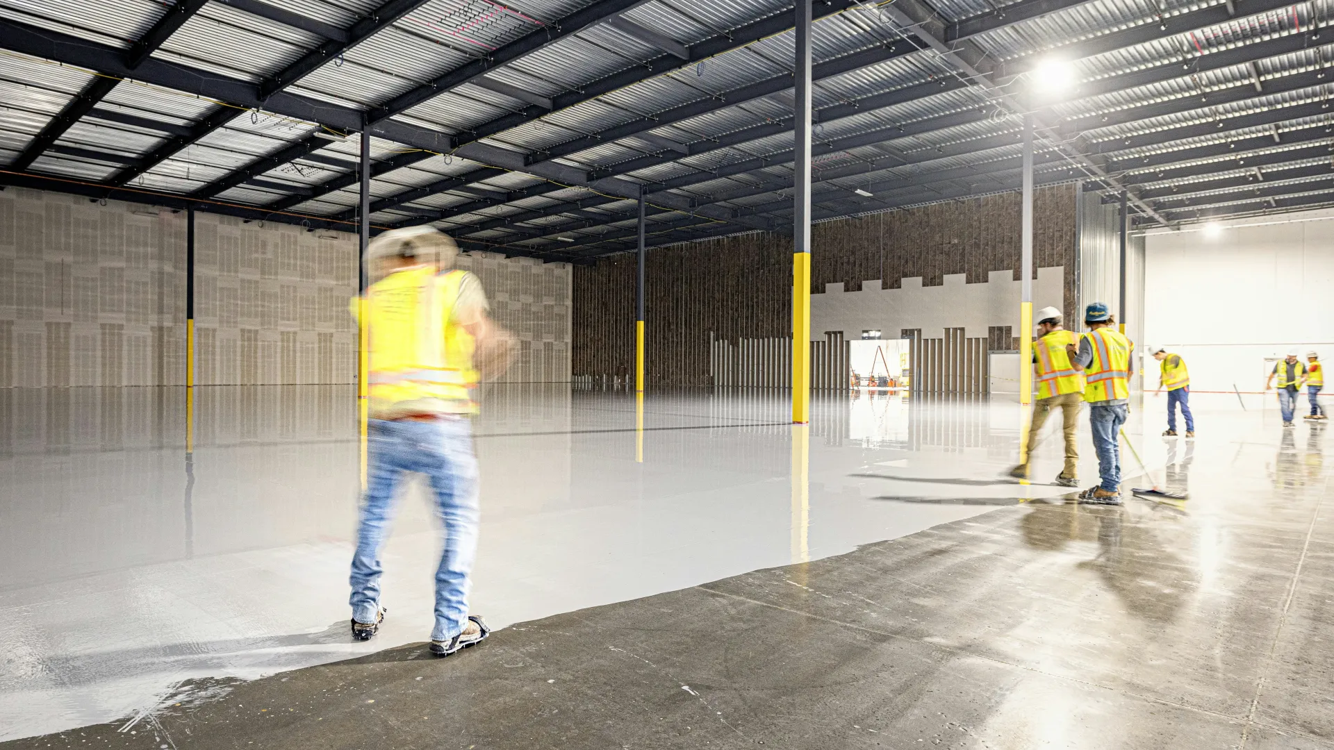 Construction workers in safety vests smoothing and finishing a large concrete floor inside a warehouse.