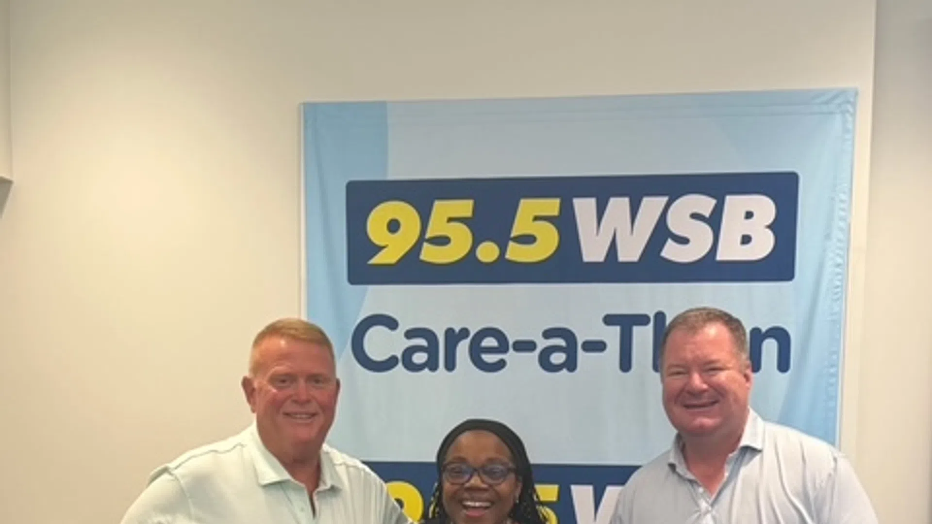 Three people smiling and posing in front of a 95.5 WSB Care-a-Thon banner in a radio station studio.