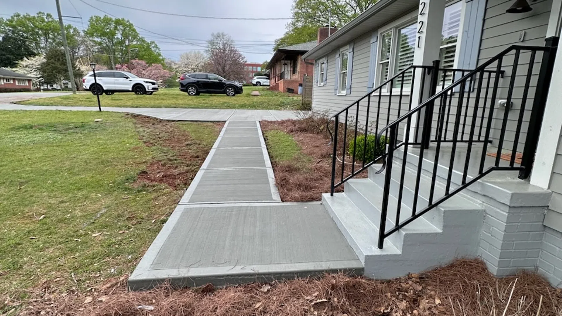 New concrete sidewalk and steps with black metal railing leading to a gray house with green lawn and parked cars in the background.