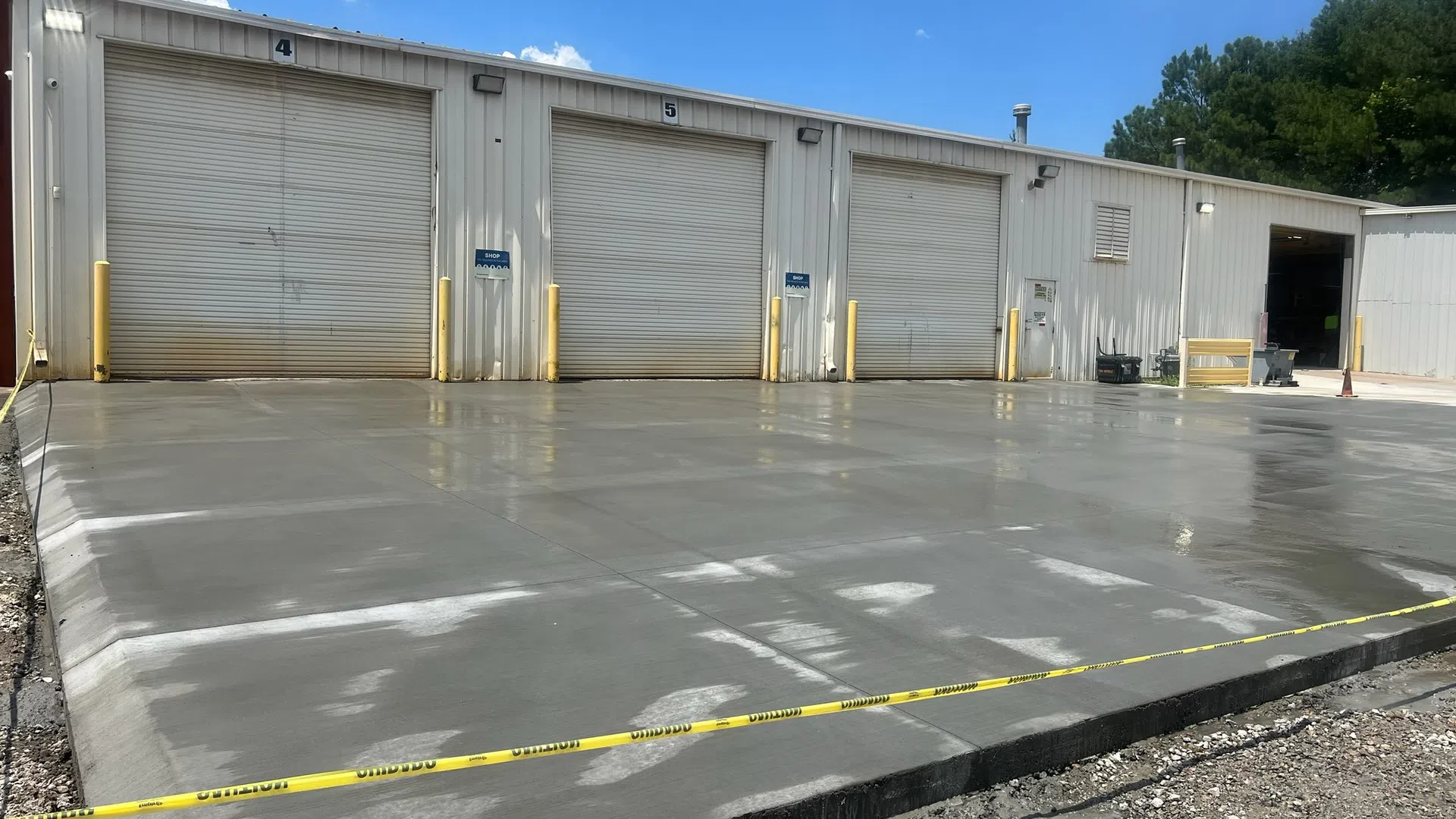 Newly poured concrete slab in front of warehouse garage doors with caution tape around the area under a blue sky.