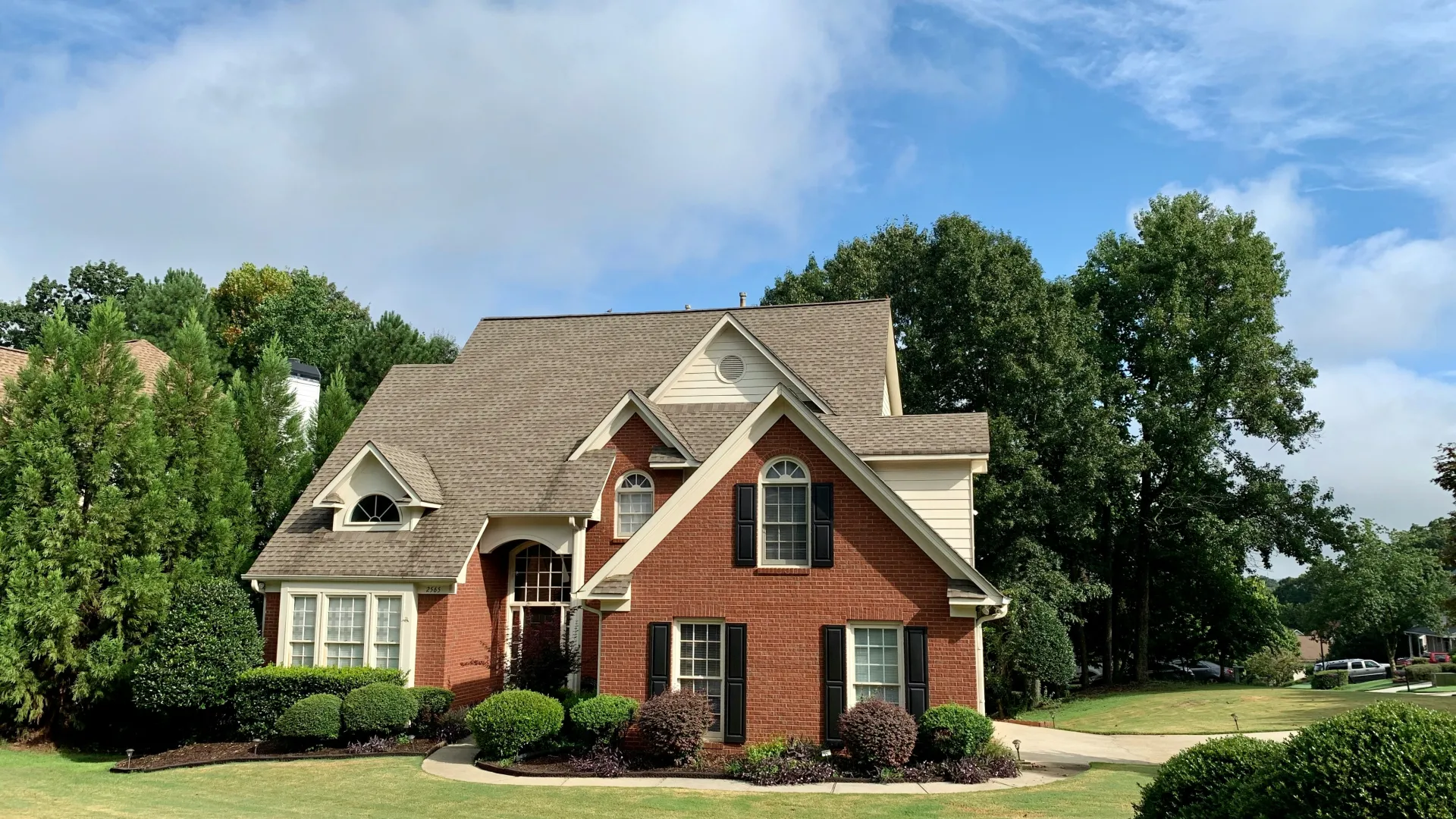 Two-story brick house with a beige roof surrounded by green trees and a neatly trimmed lawn under a blue sky.