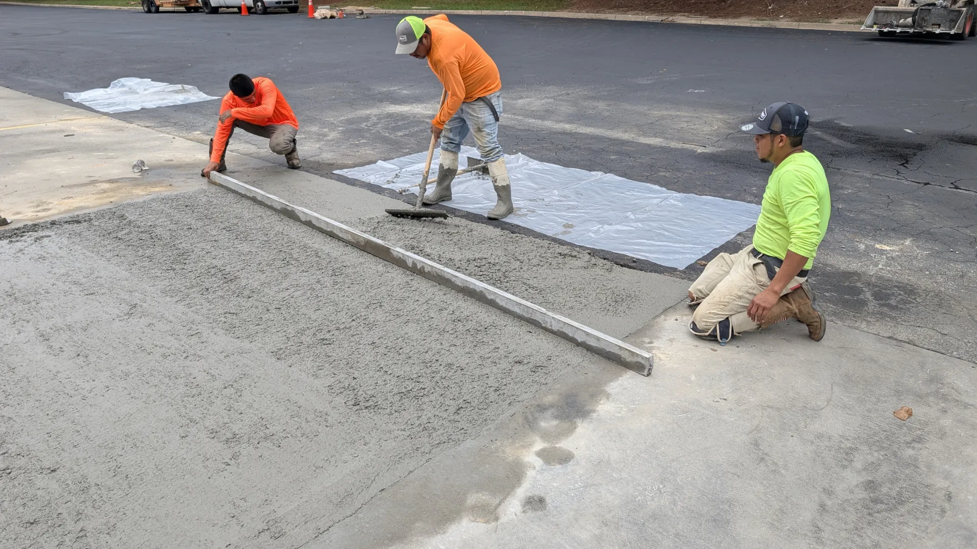 Three construction workers smoothing fresh concrete on a sidewalk near an asphalt parking lot.