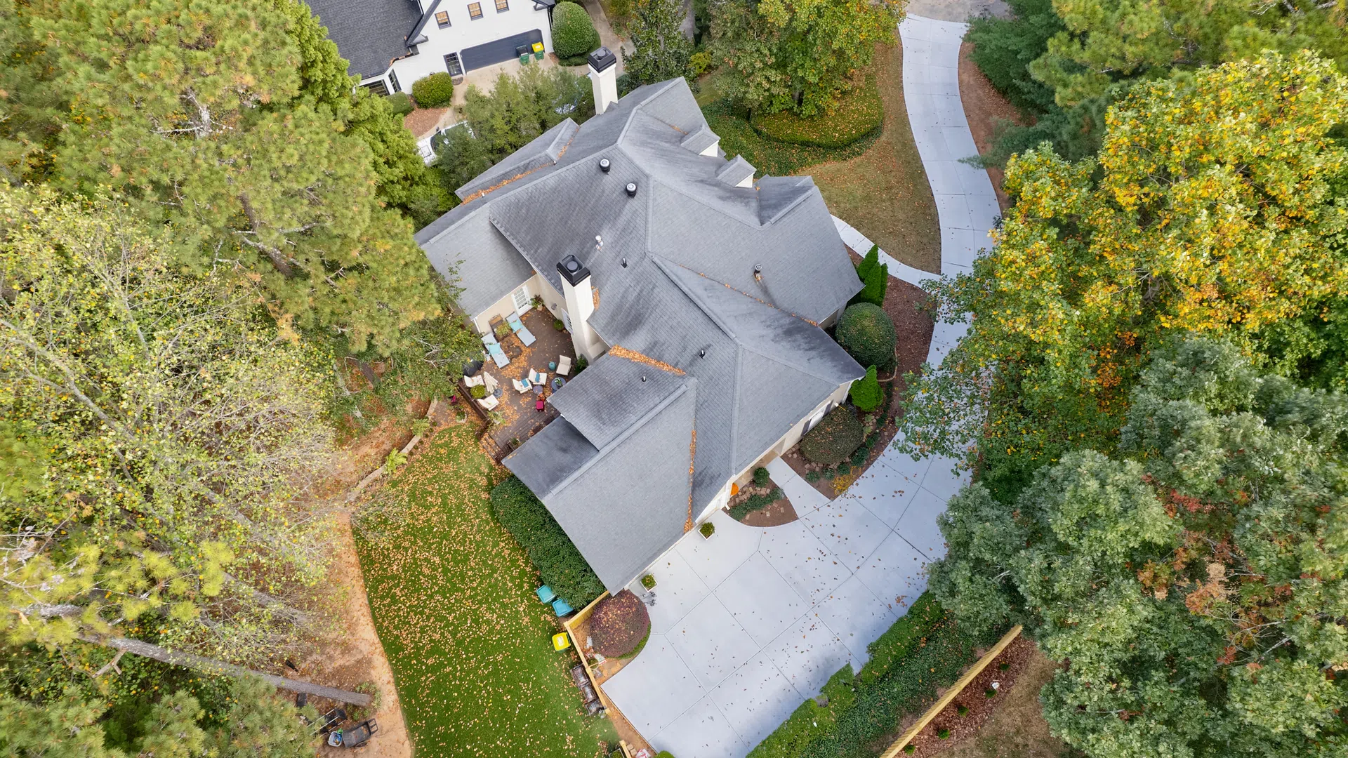 Aerial view of a suburban house with a gray roof, concrete driveway, patio, and surrounding trees in autumn.