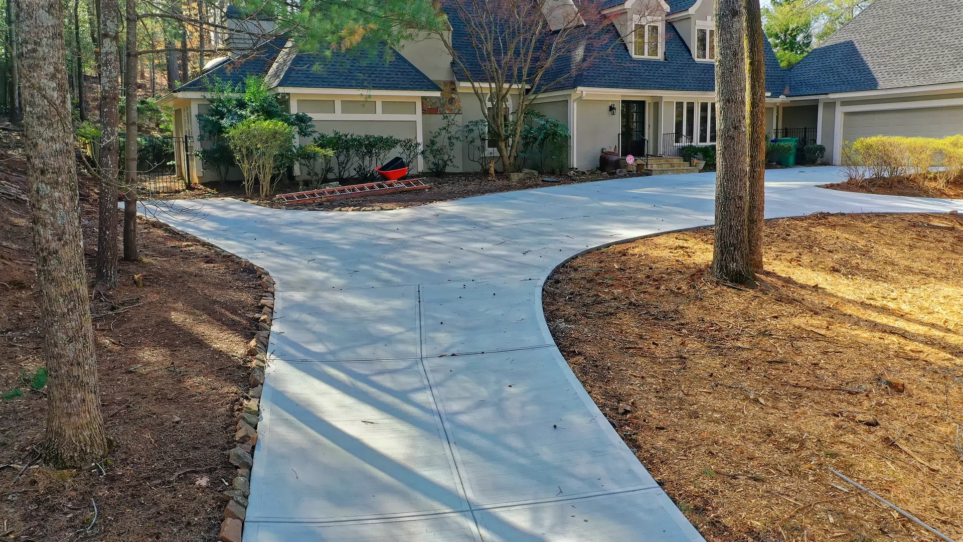 Curved concrete driveway leading to a large suburban house surrounded by trees and landscaped yard.