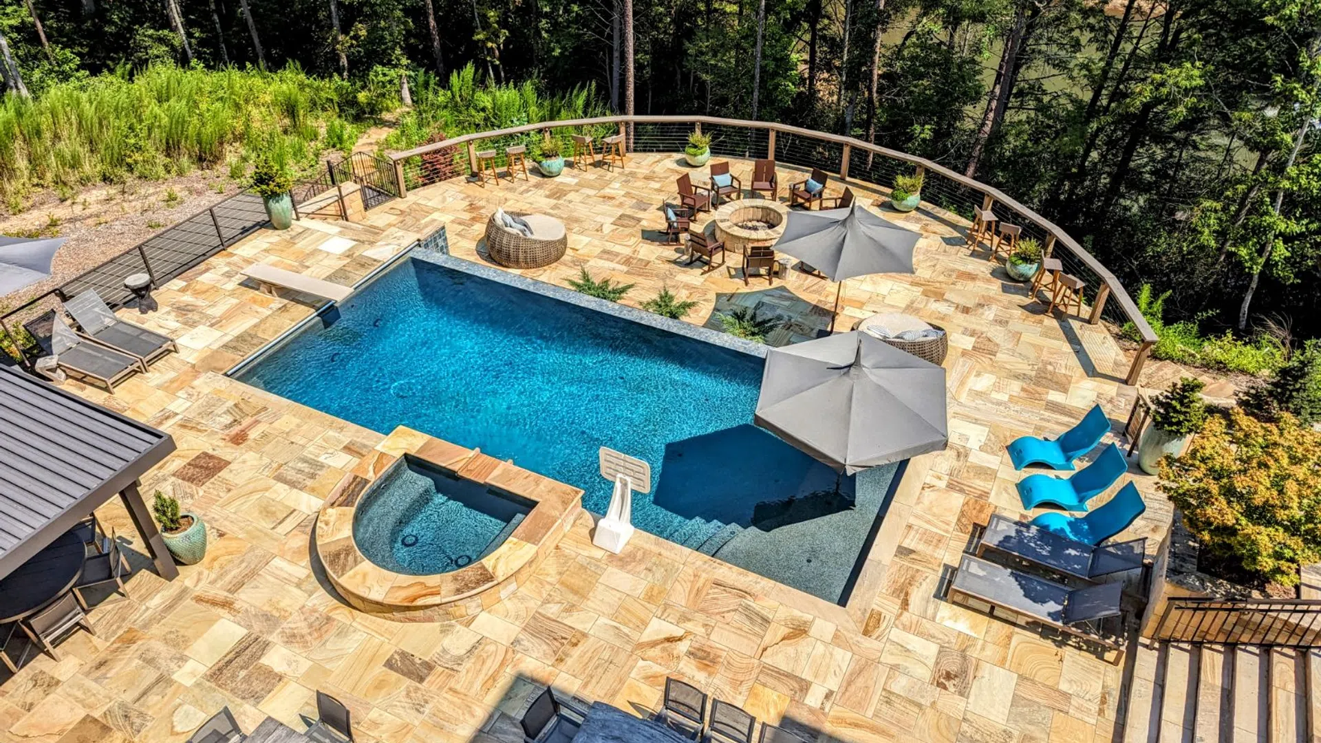 Aerial view of a backyard pool area with lounge chairs, umbrellas, fire pit, and natural stone patio surrounded by trees.