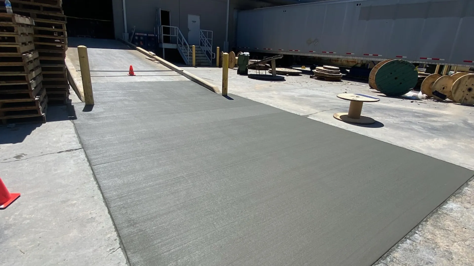 A loading dock with a new gray surface and safety cones, featuring pallets and spools in the background.