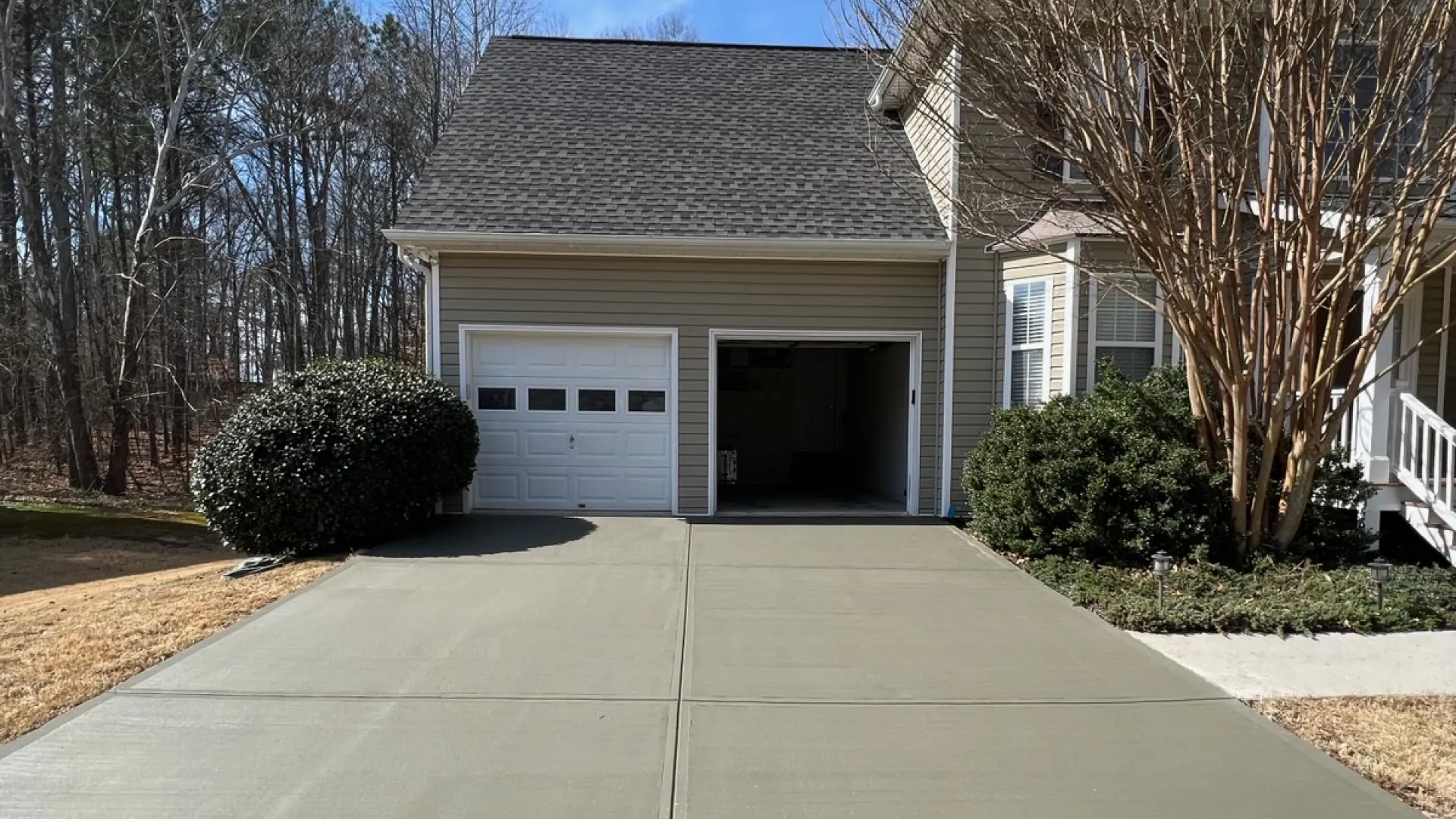 Freshly poured concrete driveway leading to a two-car garage attached to a beige house with surrounding trees and shrubs.