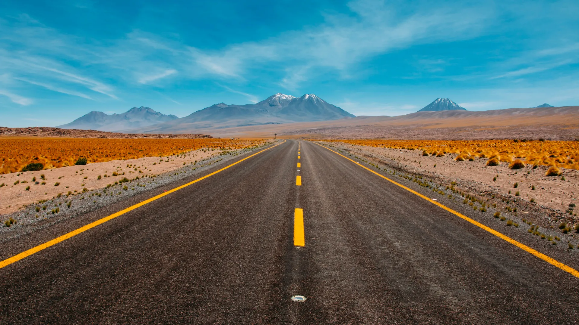 Straight pavement road with yellow lines leading to distant mountains under blue sky in arid landscape.