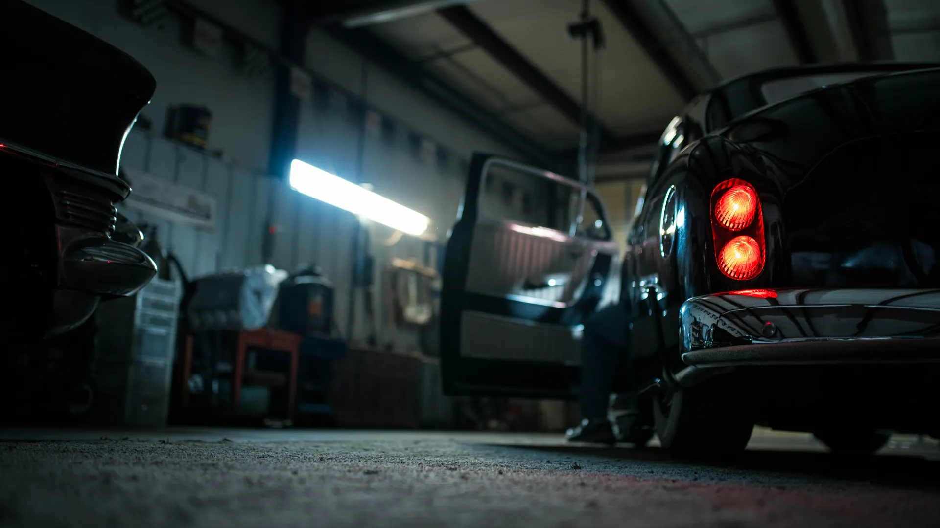 Rear view of classic black car with taillights on in dimly lit garage with open door and person inside.