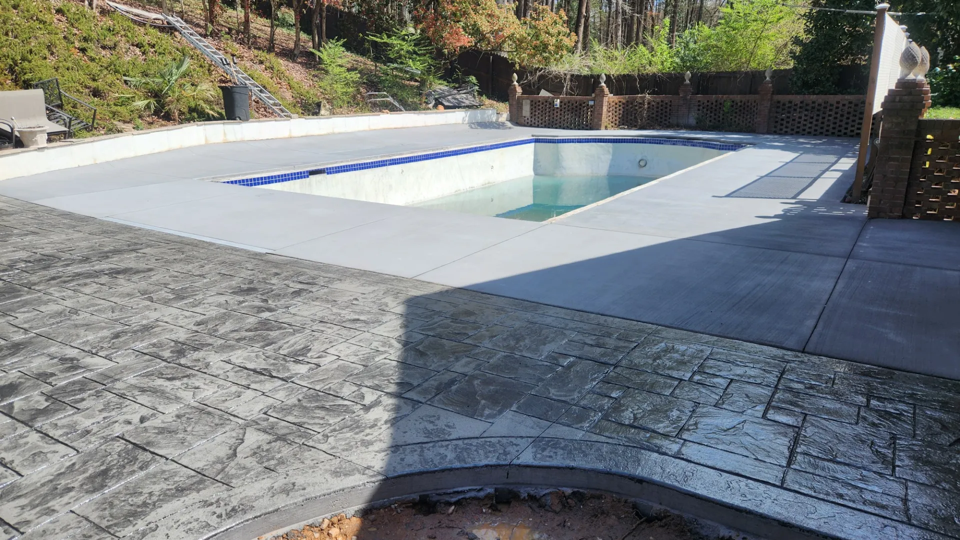 Empty rectangular swimming pool with concrete deck and stamped concrete patio in a wooded backyard.