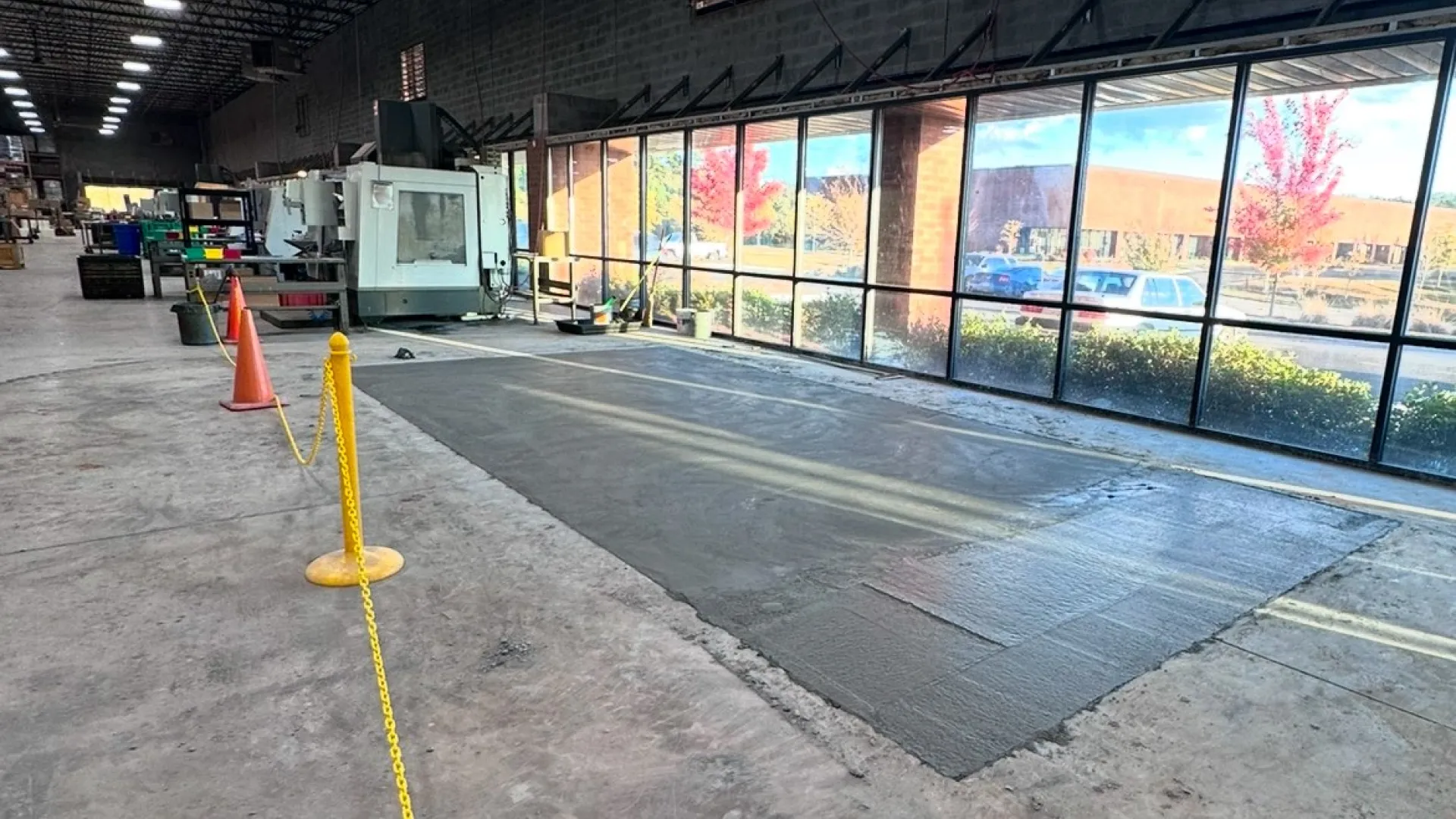 Industrial workshop with large windows, machinery, concrete floor, safety cones, and yellow chain barrier inside a warehouse.