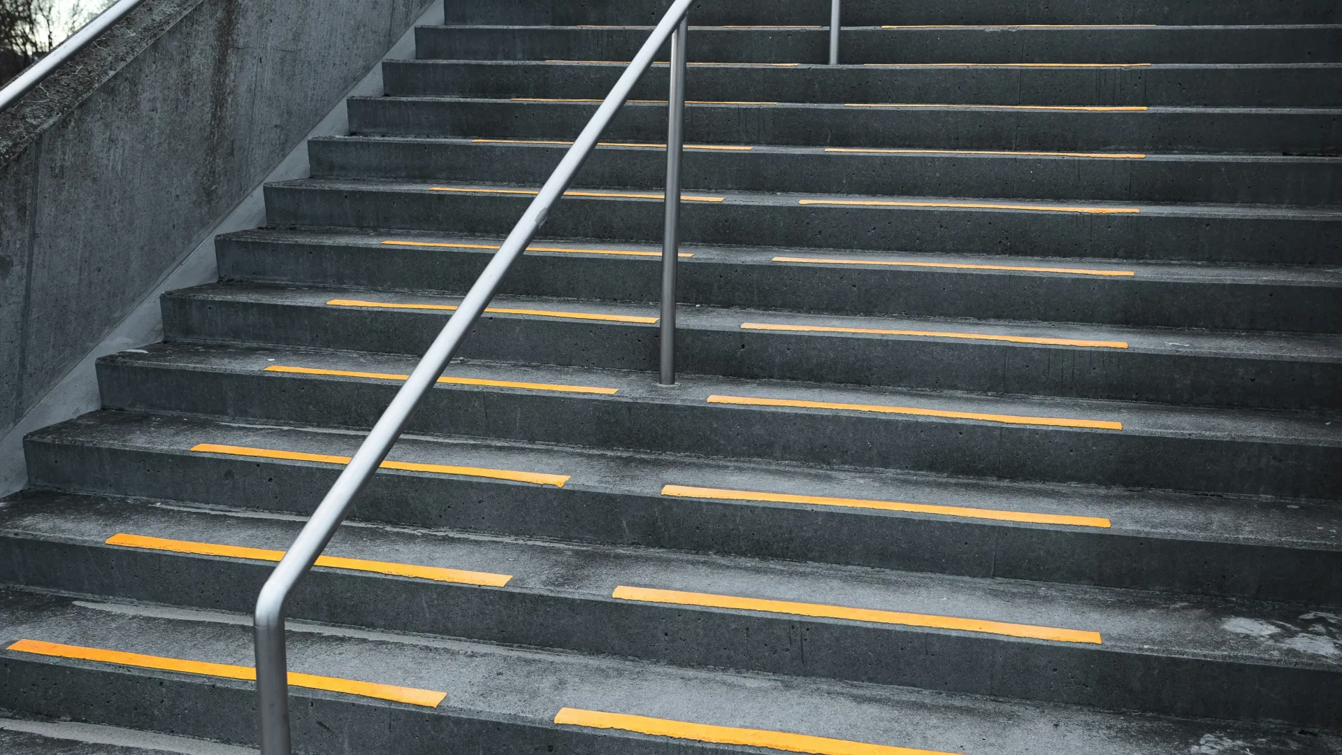Concrete outdoor stairs with yellow safety strips and a silver handrail under an overcast sky.