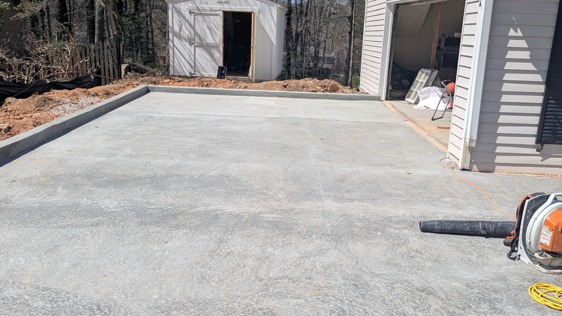 Freshly poured concrete driveway next to a white garage and a small white storage shed surrounded by trees.