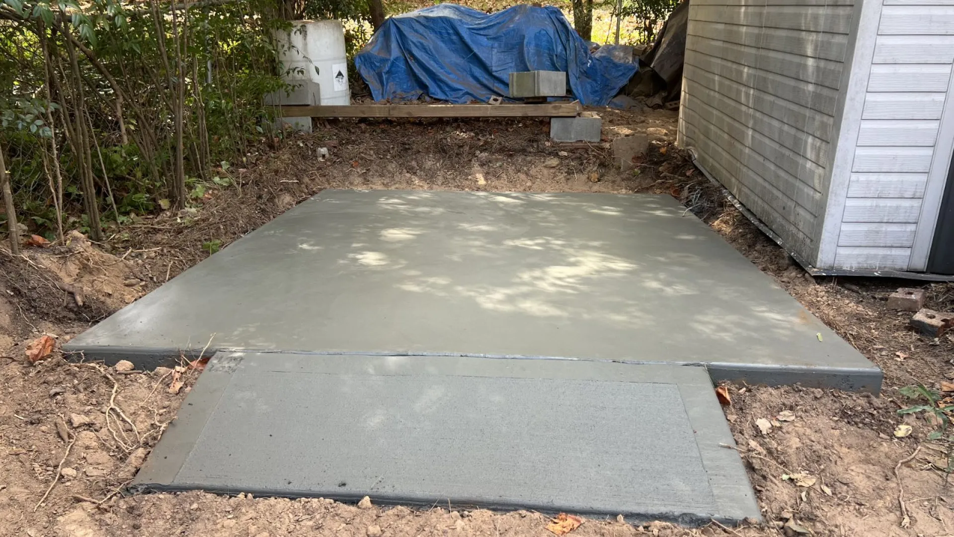 Newly poured smooth concrete slab next to a white shed in a backyard setting with trees and a blue tarp.