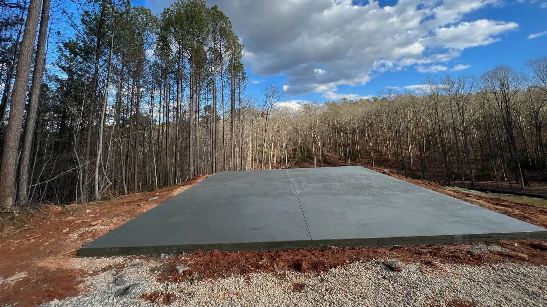 New concrete foundation slab in clearing surrounded by bare trees and a cloudy blue sky.