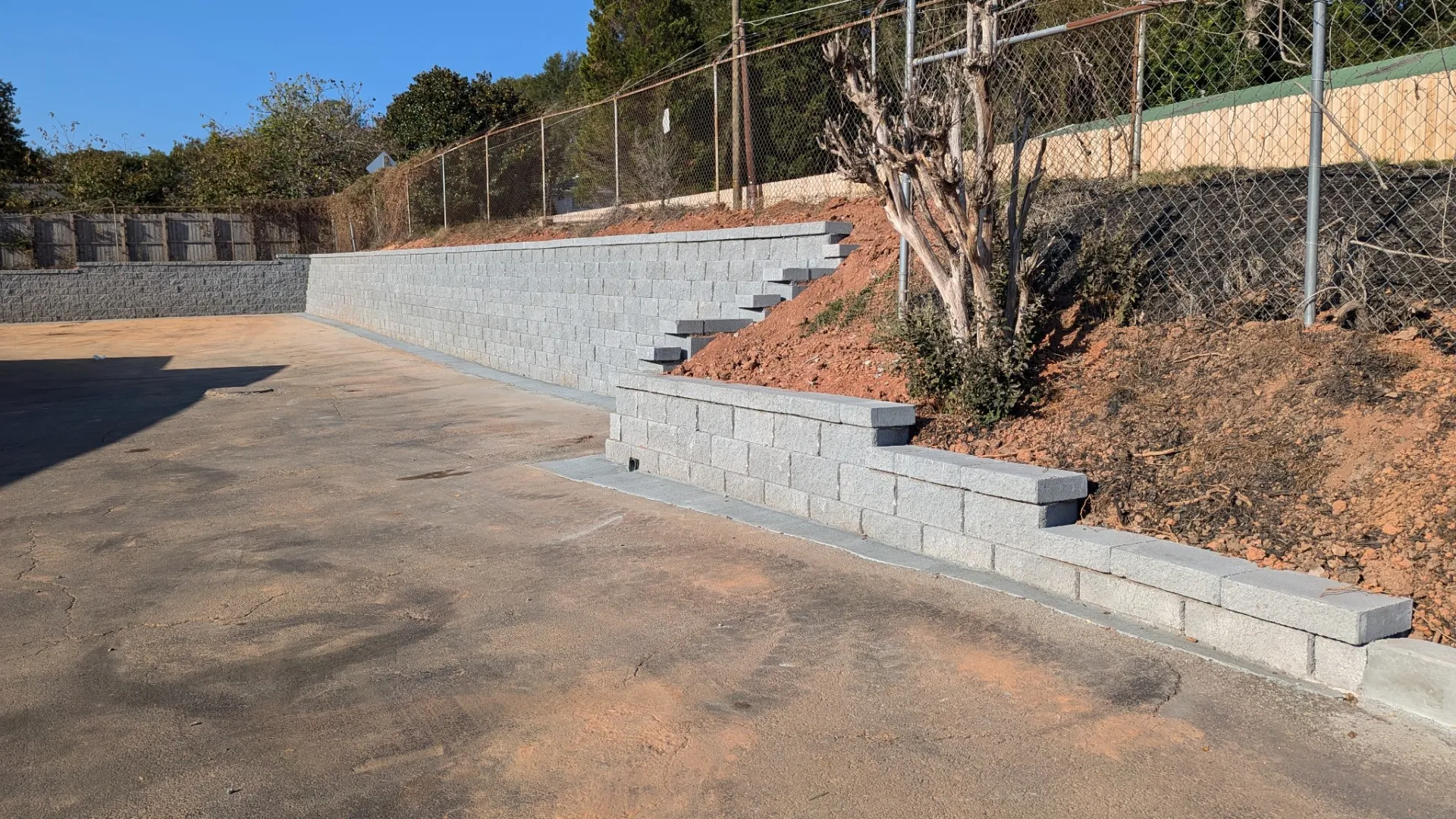 Concrete retaining walls with steps lining a dirt slope beside an asphalt area under clear blue sky.