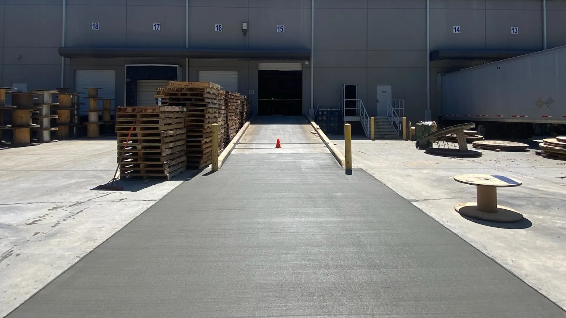 Newly poured concrete ramp leading to warehouse loading docks with wooden pallets and industrial materials around.