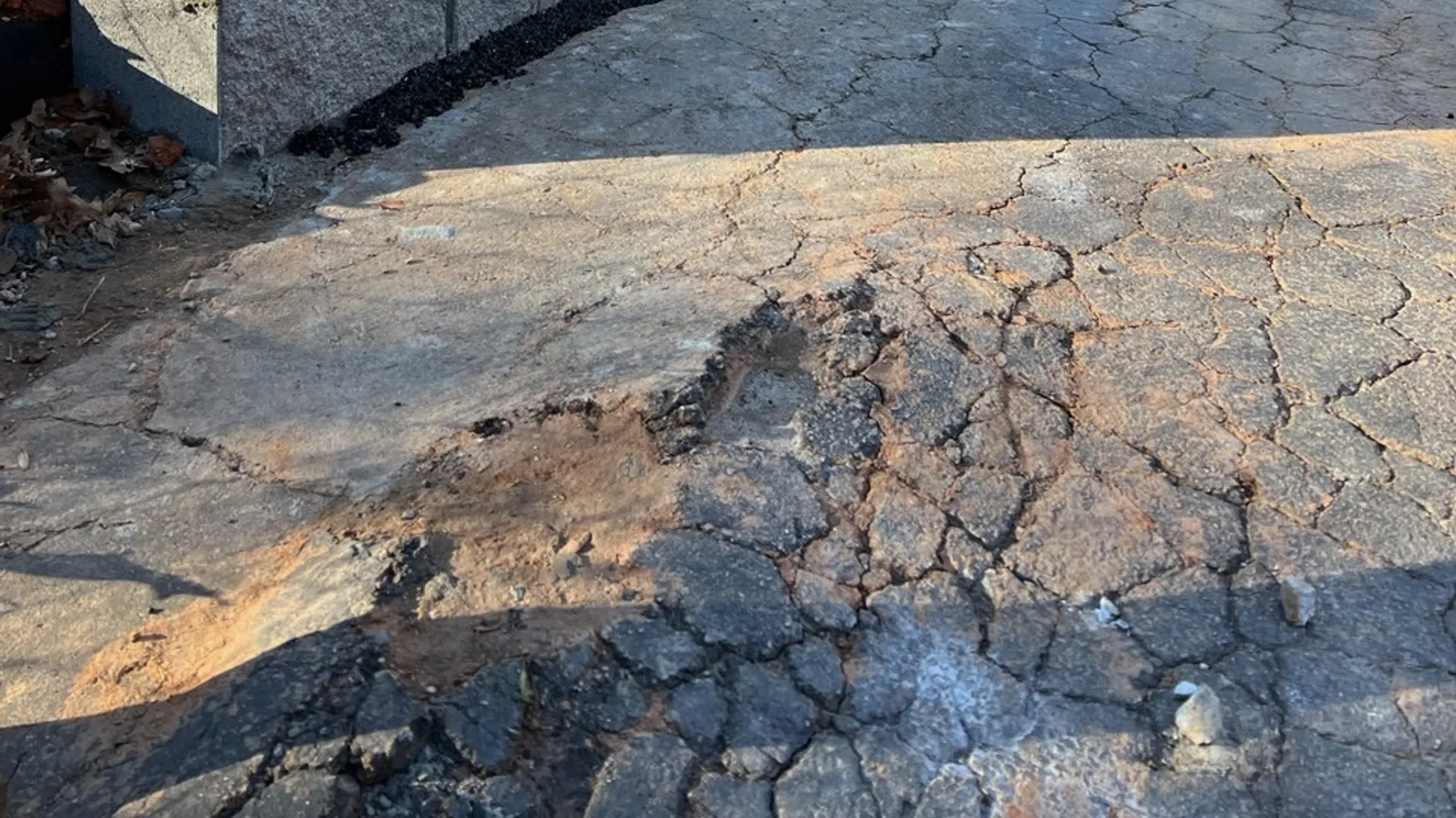 Concrete retaining wall next to cracked asphalt driveway under clear blue sky in a wooded area.