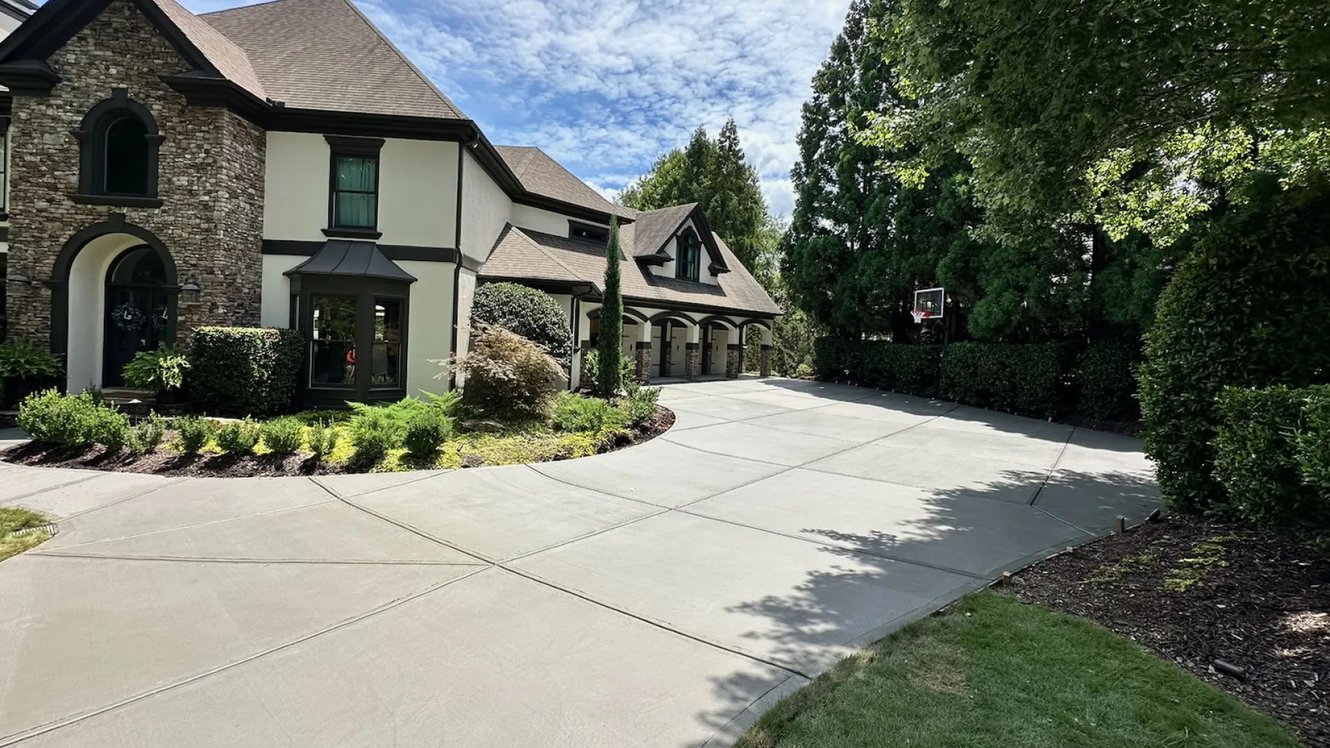 Elegant home with stone facade, lush greenery, and a curved driveway on a sunny day.
