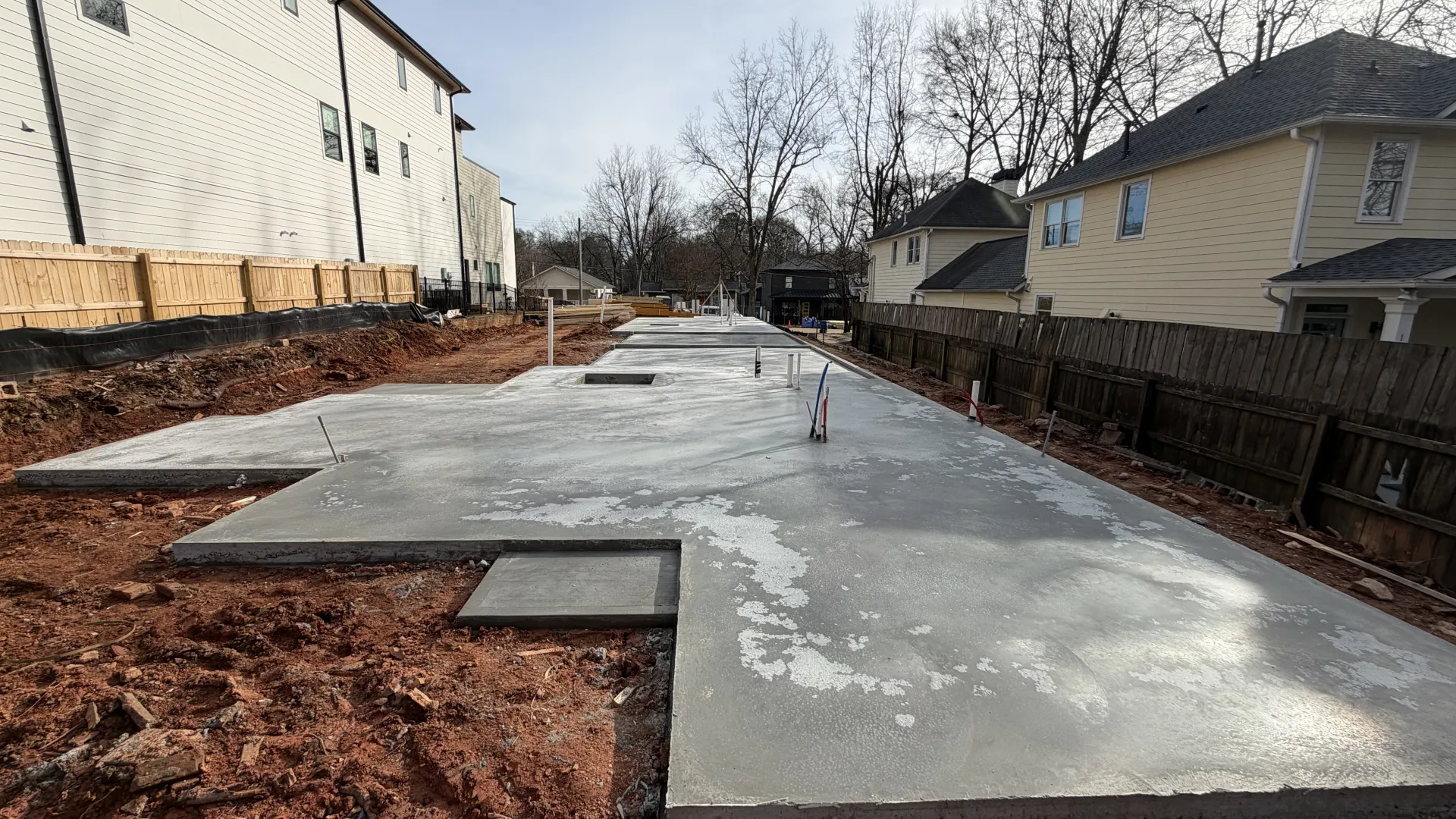 Freshly poured concrete foundation slab for a residential building surrounded by soil and nearby houses.