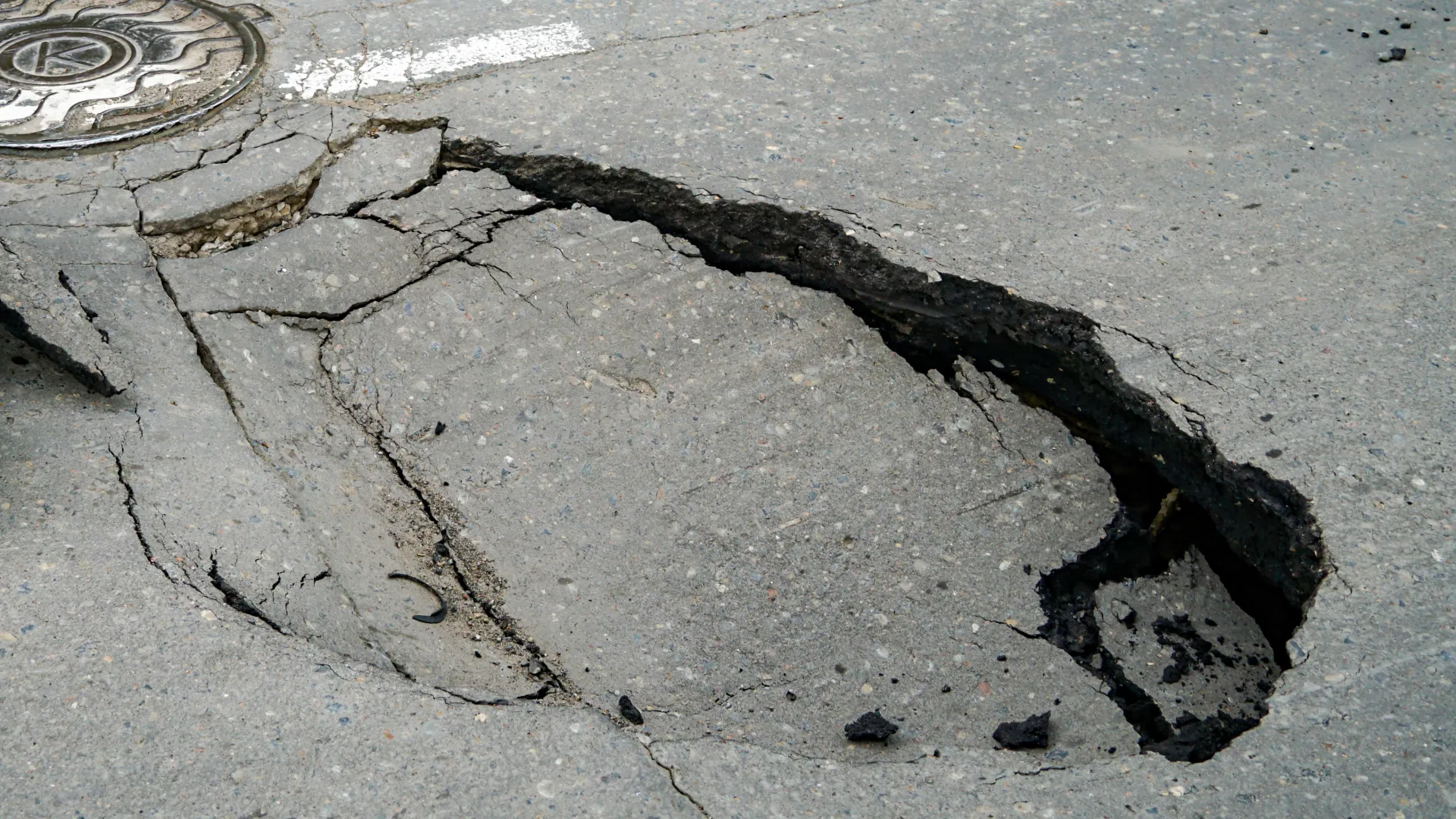 Cracked pavement with a large pothole and visible road markings, highlighting urban infrastructure issues.