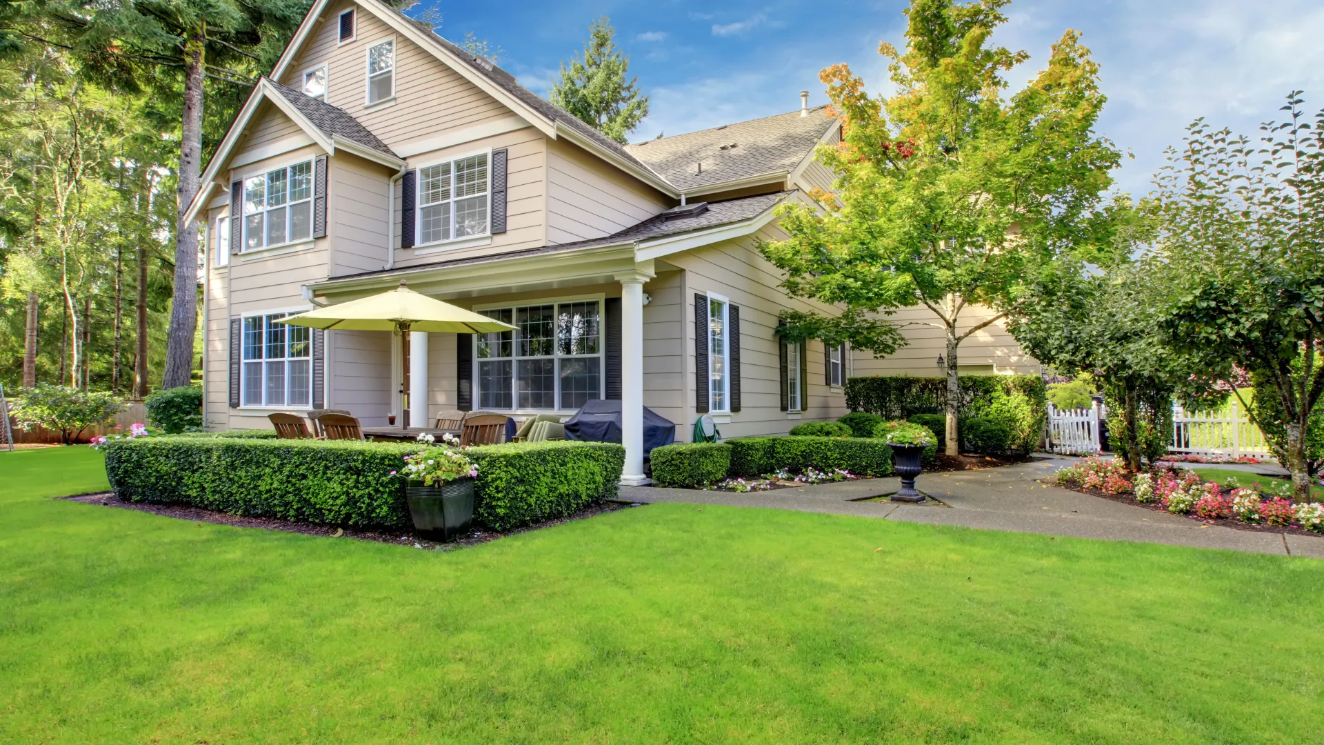 Two-story suburban house with manicured lawn, patio furniture, green umbrella, and surrounding trees on a sunny day