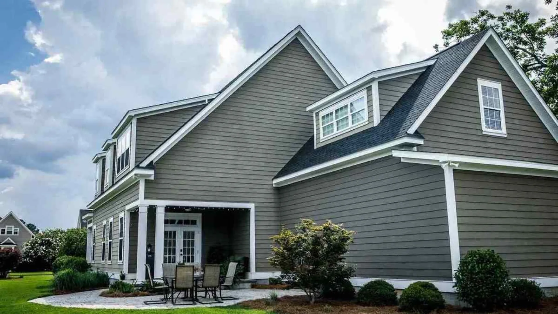 Modern gray two-story house with white trim, patio furniture, and well-maintained landscaping under a cloudy sky.