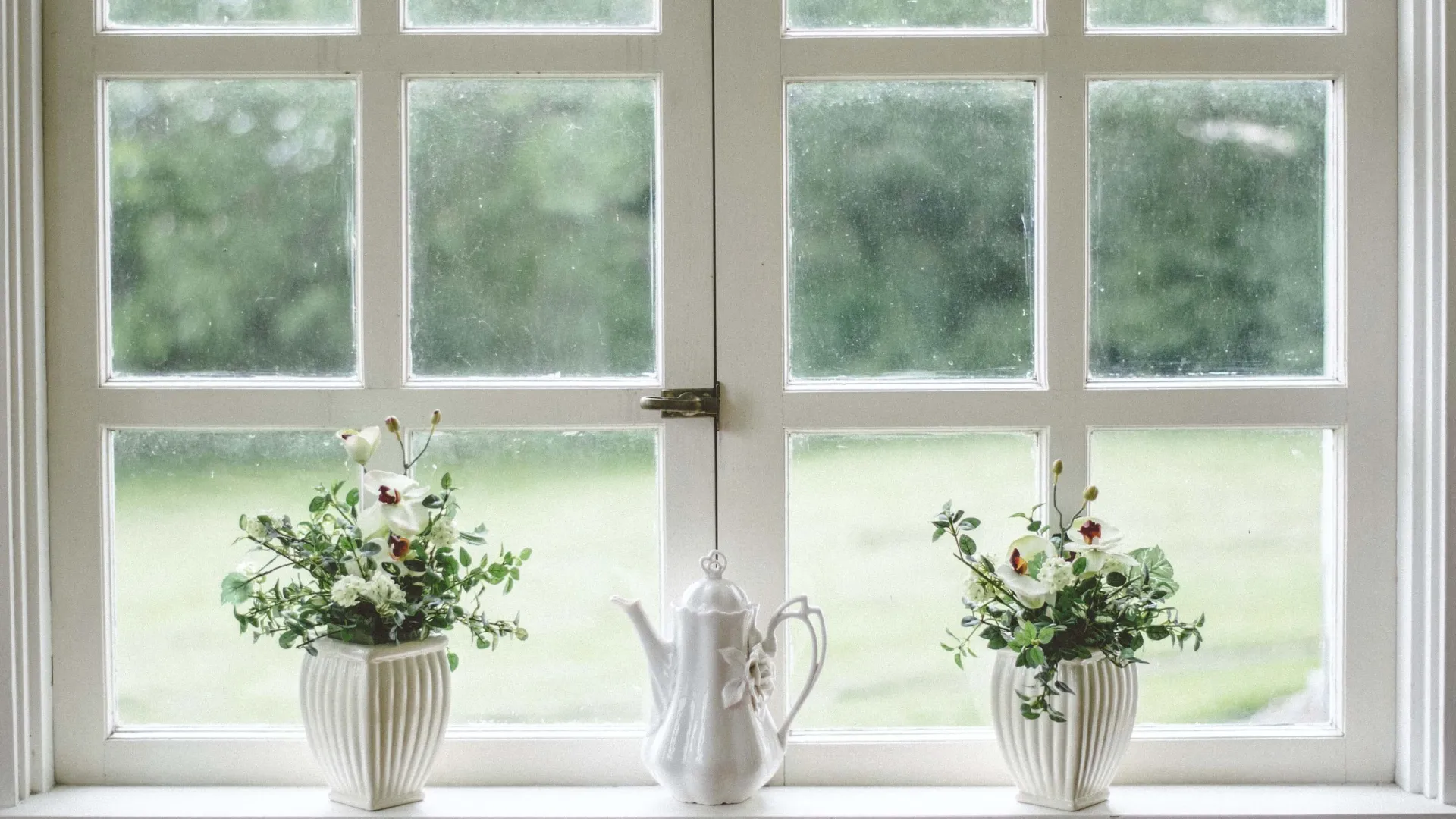 White teapot and two flower-filled white vases on a windowsill with green outdoors visible outside.