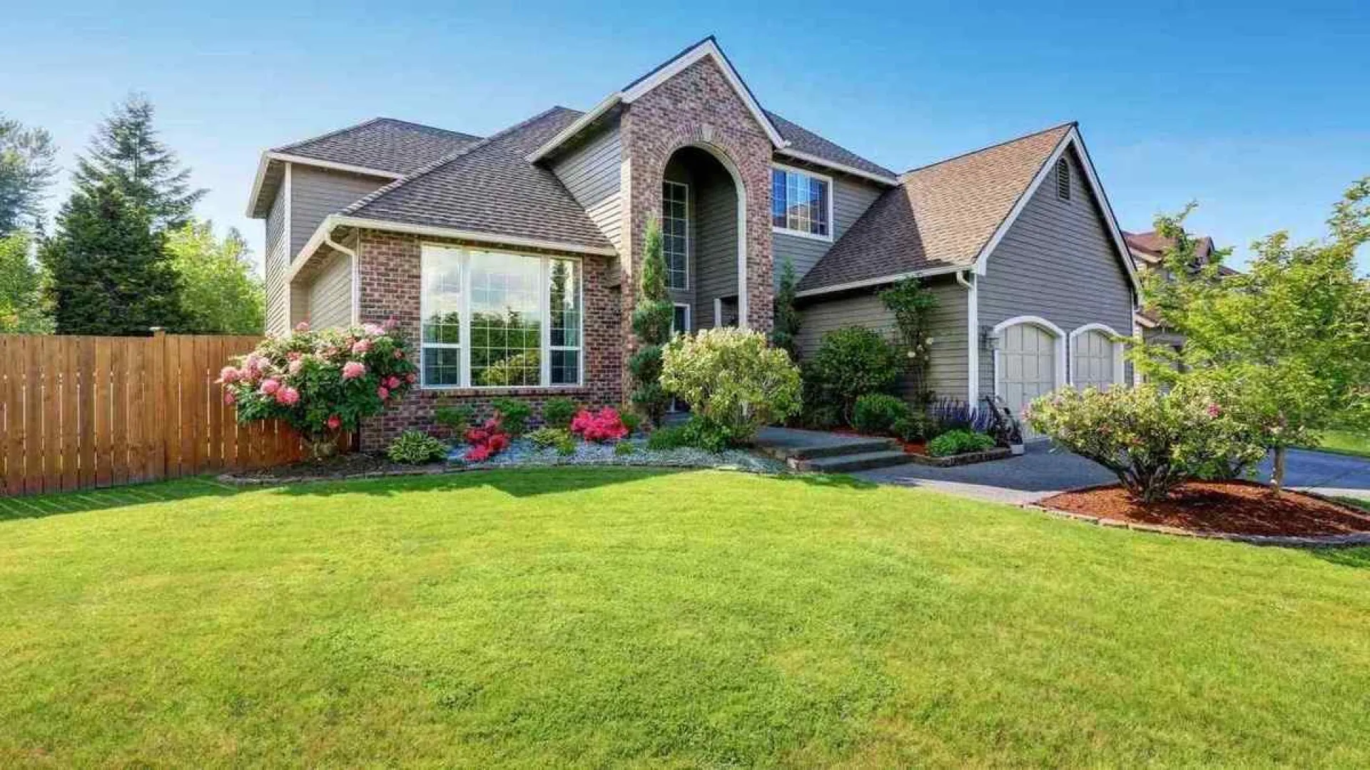 Suburban house with brick and siding exterior, well-manicured lawn and colorful flower beds under clear blue sky