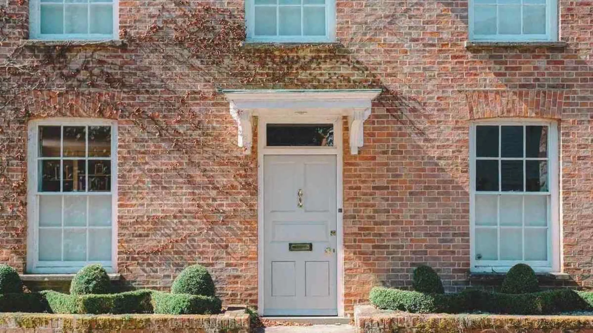 Traditional red brick house with white front door and symmetrical windows surrounded by manicured green shrubs