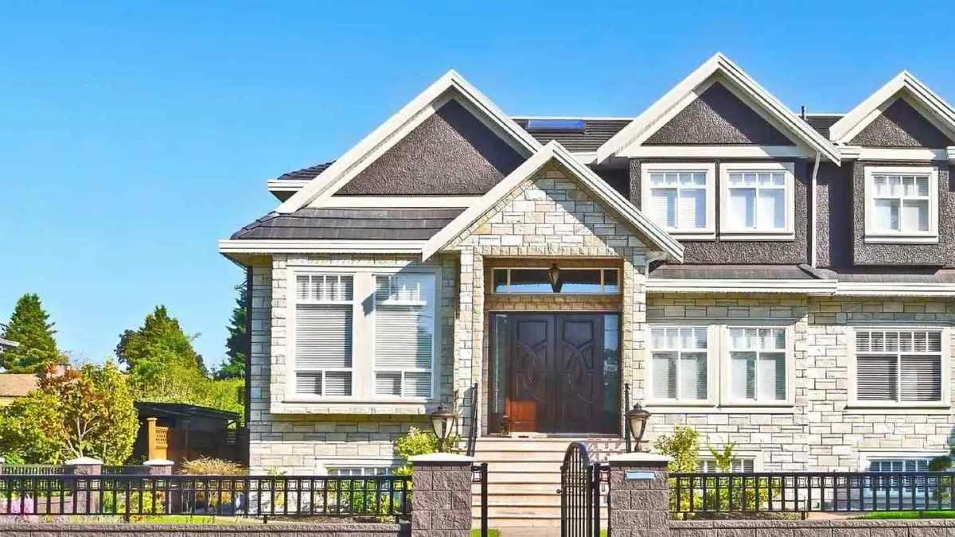 Modern two-story stone house with large windows, double front doors, triangular roof, and black iron fence.
