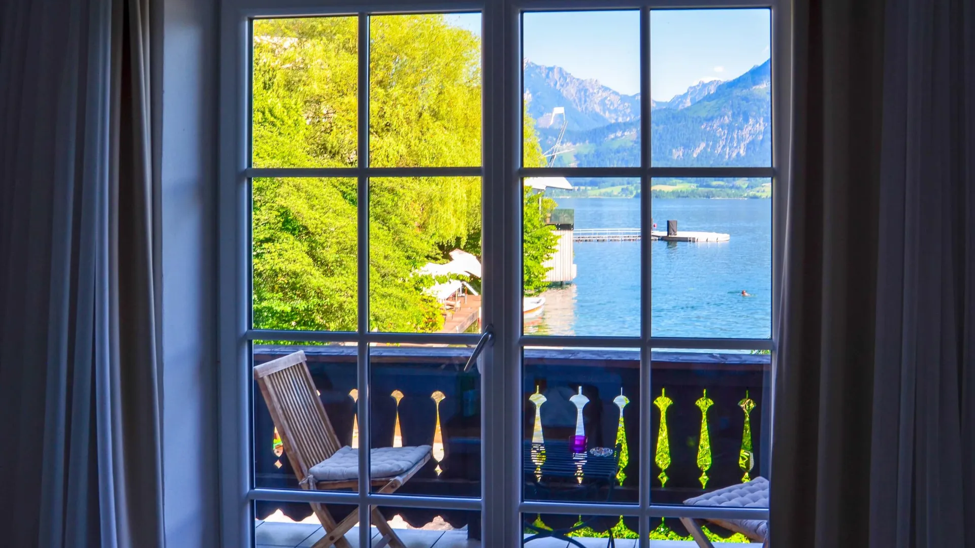 View through glass doors of wooden chairs on balcony overlooking lake, mountains, and lush green trees on a sunny day