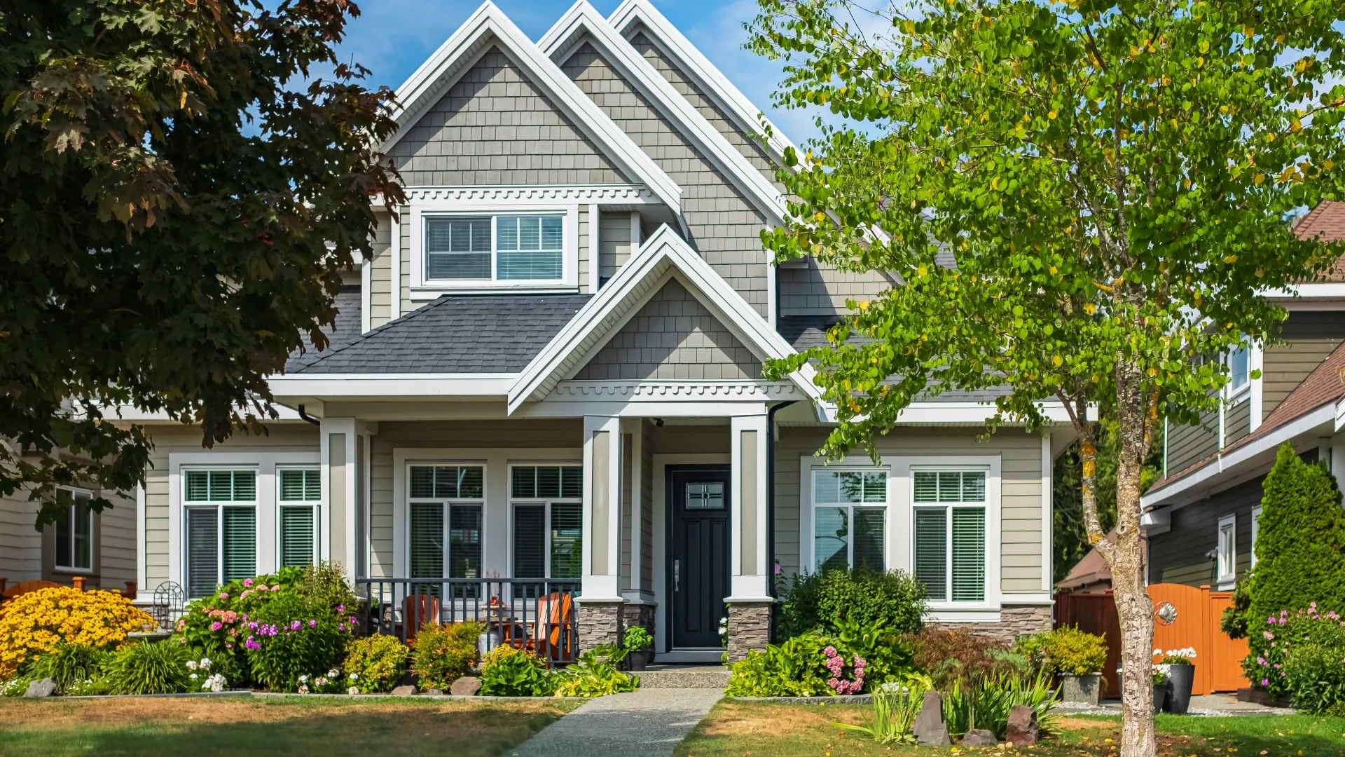 Modern two-story house with gray siding, white trim, lush landscaping, and a front porch under a blue sky.